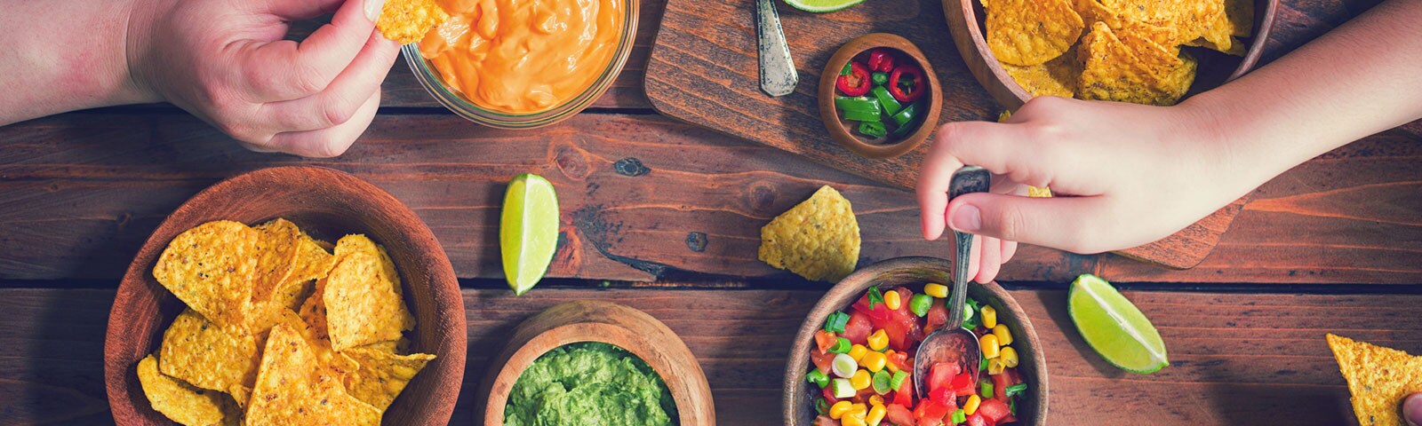 On a wooden table sits bowls of queso, salsa, guacamole and corn chips with hands reaching in to grab a serving