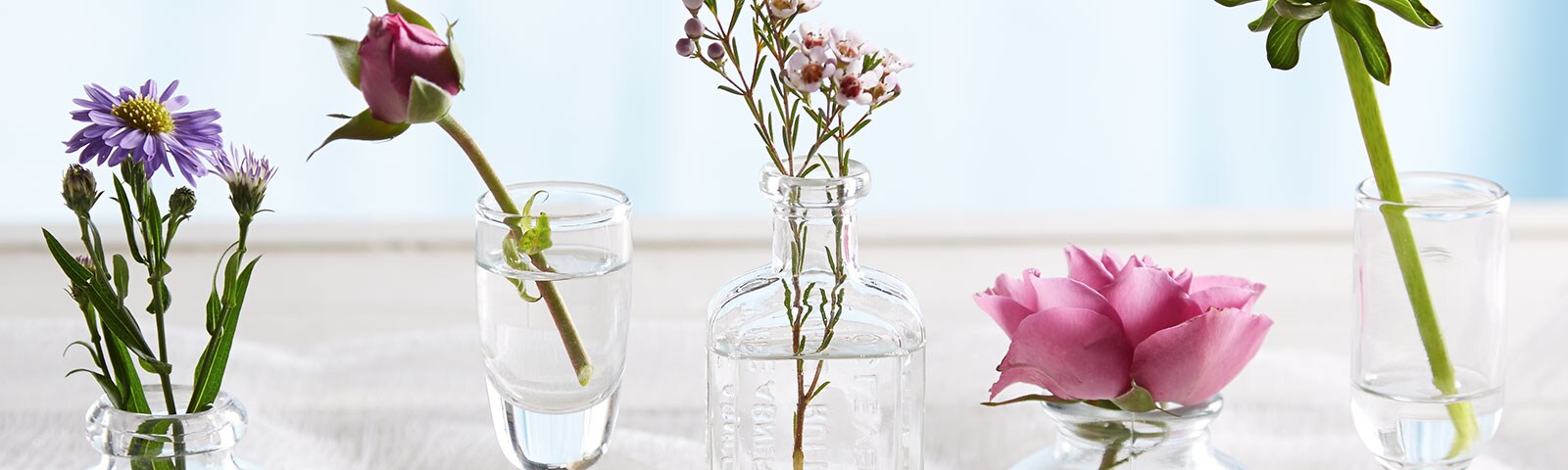 Five glass containers of various shapes and size, each containing water and a different flower 