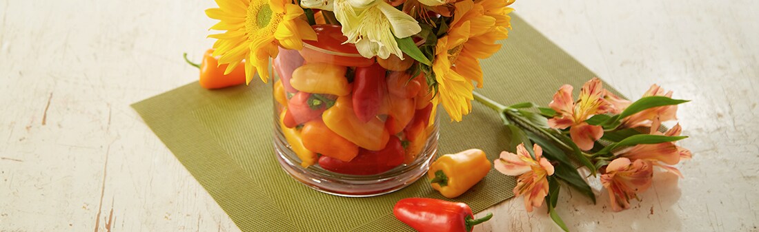 A vase full of multicolored mini peppers with sunflowers coming out of the top