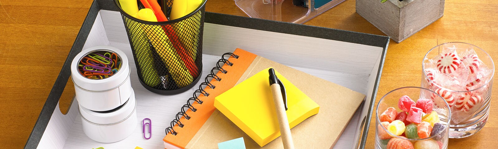 Homemade desk organizer with notepads, pens and paperclips inside next to candy-filled cups on a wooden table