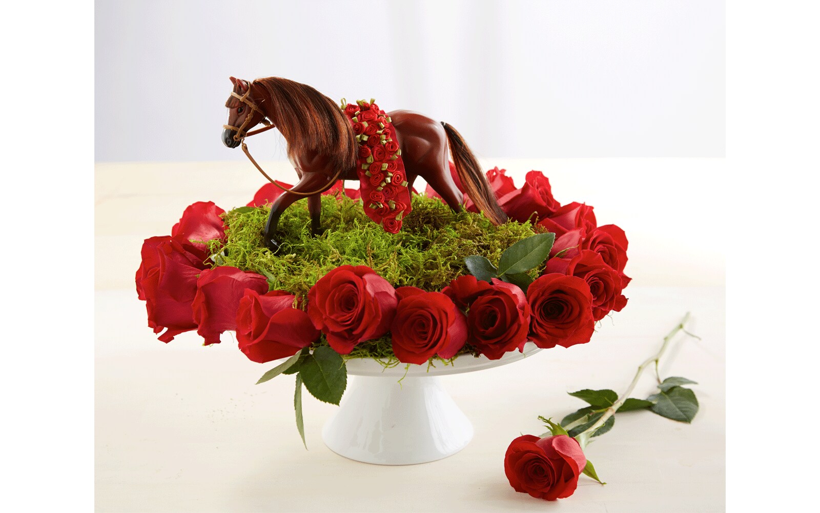A small horse figurine with a sash of roses on a field of fake grass sits atop a cake plate adorned with roses