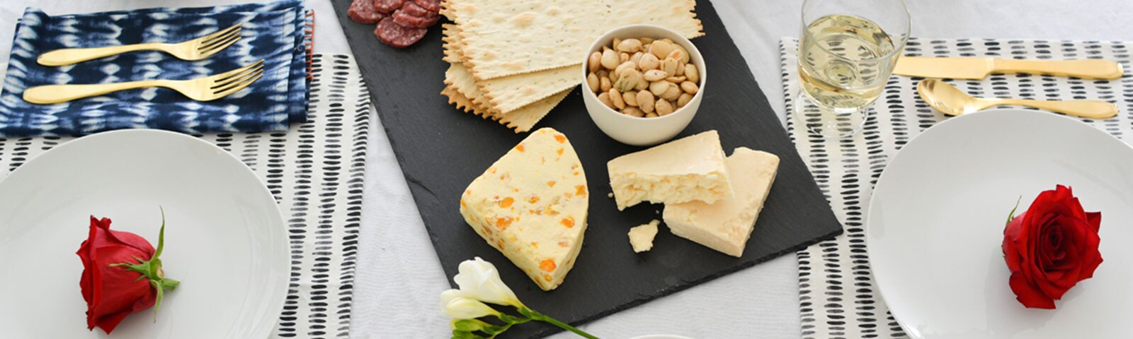 A table on top of which lays a charcuterie board, linen napkins with golden forks, and round white plates with a rose flower in the center