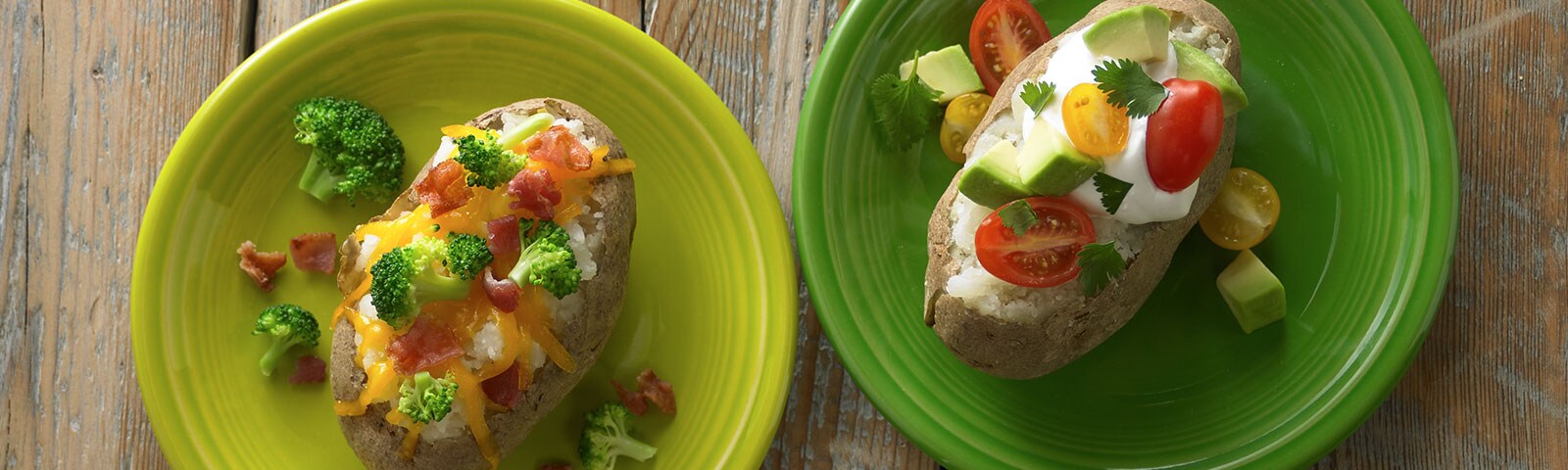 Two baked potatoes sitting on separate plates on a wooden table. Each potato is loaded with various toppings from bacon, to broccoli, to avocado.