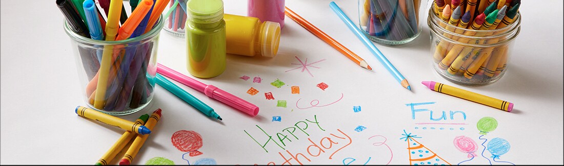 On a paper table cloth sits markers, crayons and other craft supplies with Happy Birthday written on the paper in the middle