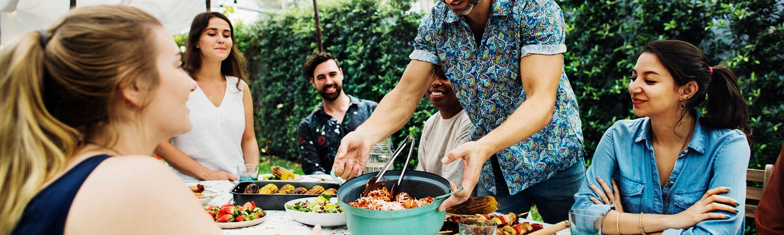Party guests seated around a table smiling while a man lowers a bowl of food onto the center of the table. 