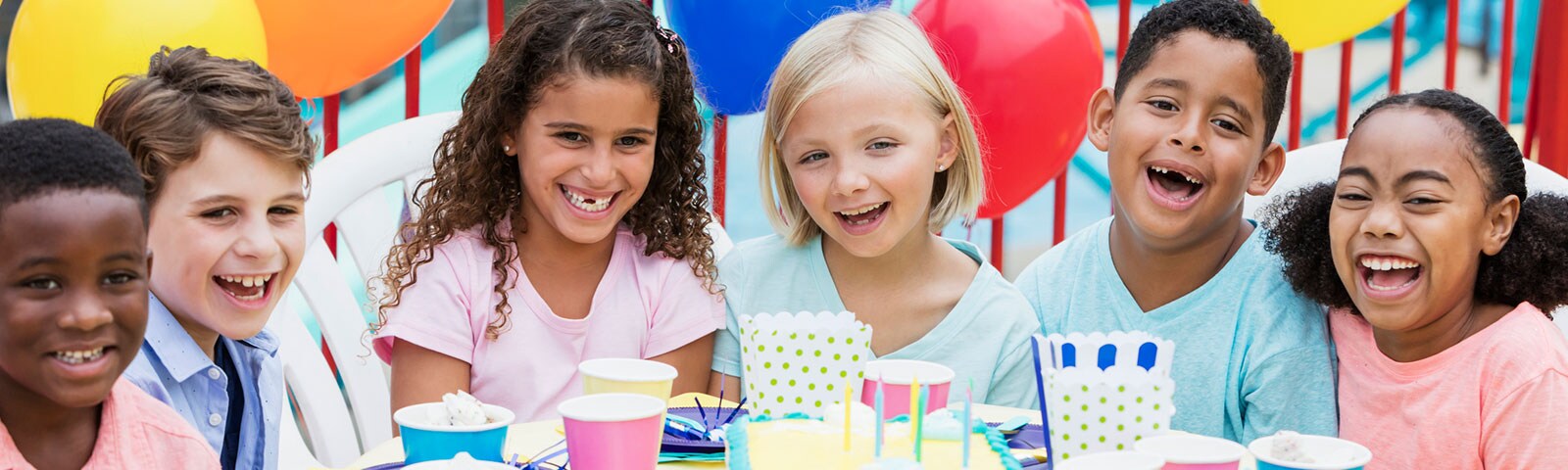 Six kids laughing while sitting around a table with a birthday cake in the middle