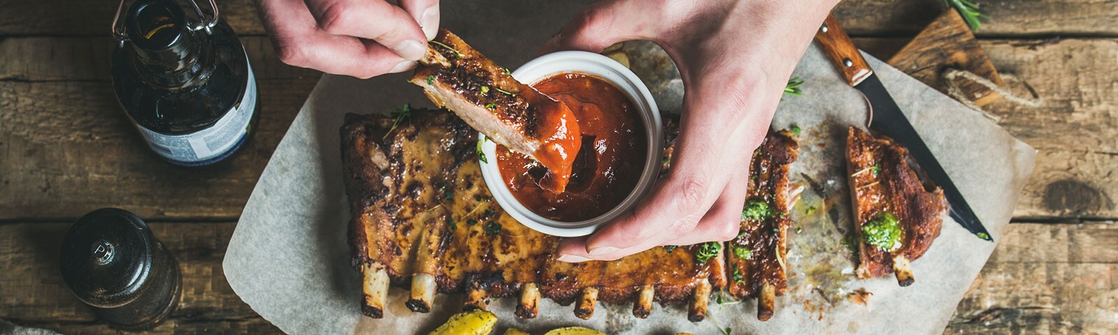 A pair of hands dipping beef ribs into barbecue sauce